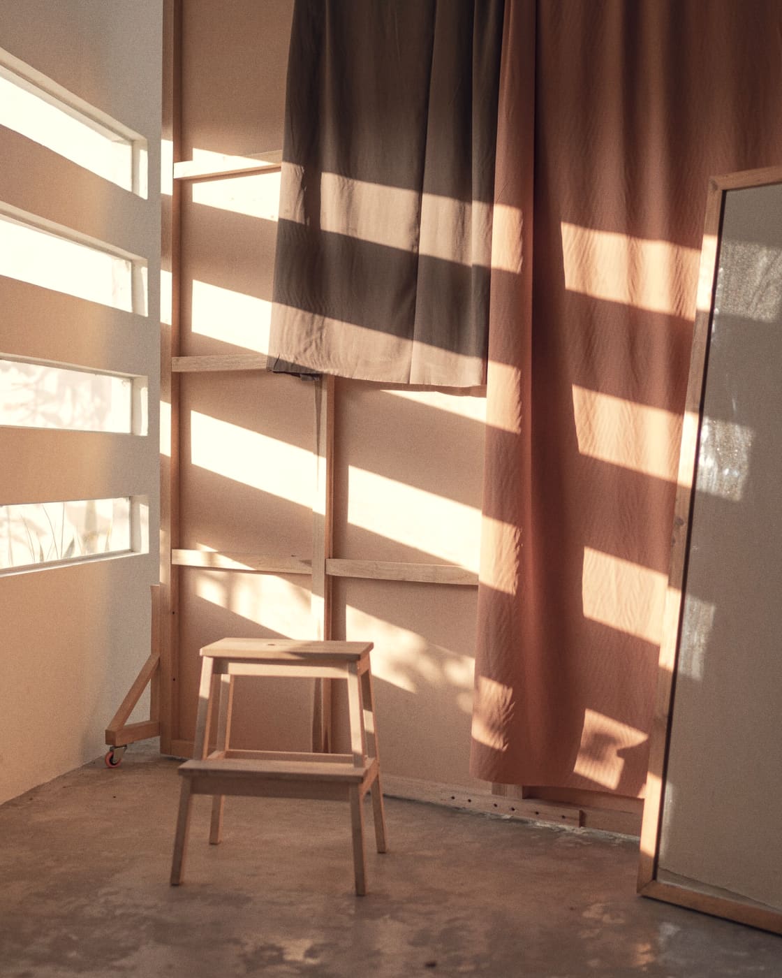 An empty studio room at early morning, sunlight falling in bars across a plaster wall and a warm curtain